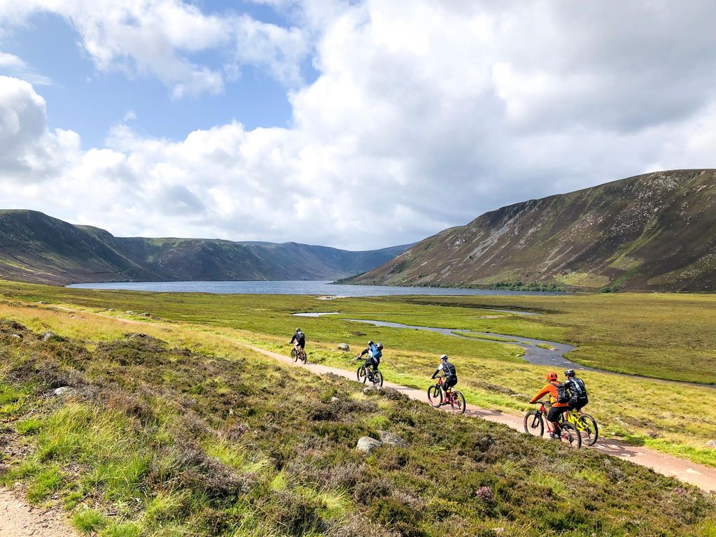 Group of people riding bicycles near a lake, lochnagar, during daytime, mountains appear in the background.